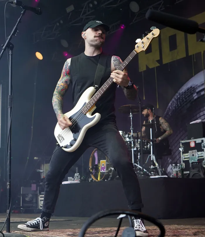 Ein tätowierter Musiker in schwarzem ärmellosem Shirt spielt einen weißen E-Bass auf einer Bühne.