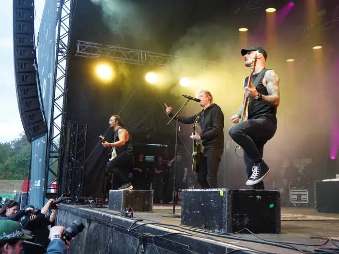 Die Band Rogers spielt auf einer hell erleuchteten Bühne mit Bühnennebel, wobei ein Bassist im Vordergrund steht.
