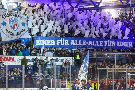 Eishockeyfans jubeln auf der Tribüne und halten ein Banner mit der Aufschrift „Einer für alle – alle für einen“ hoch. Im Vordergrund die Spieler der Iserlohn Roosters auf dem Eis.