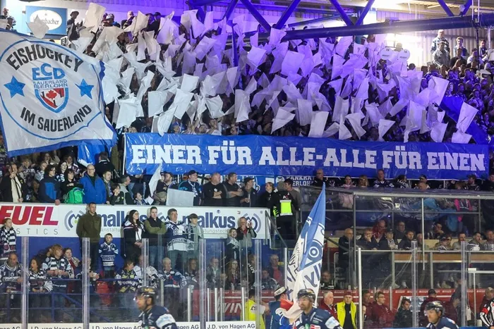 Eishockeyfans jubeln auf der Tribüne und halten ein Banner mit der Aufschrift „Einer für alle – alle für einen“ hoch. Im Vordergrund die Spieler der Iserlohn Roosters auf dem Eis.