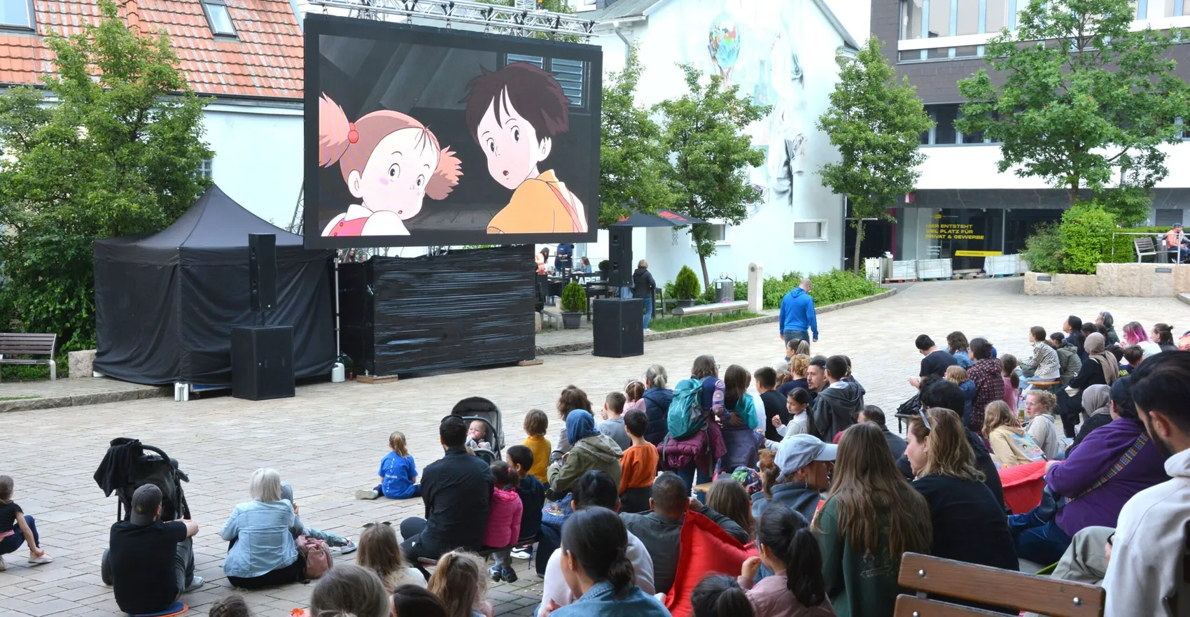 Zuschauer sitzen auf Decken und Stühlen vor einer großen Leinwand, auf der ein Anime-Film läuft. Im Hintergrund sind Gebäude und Bäume zu sehen.