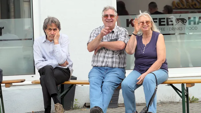 Drei Personen sitzen auf einer Bank vor einem Café; ein Mann im karierten Hemd und eine Frau im lila Oberteil lächeln.