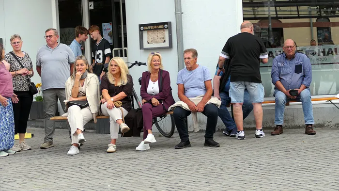 Eine Gruppe von Menschen sitzt auf einer Bank vor einem Gebäude mit der Aufschrift „Kaffee Haus“.