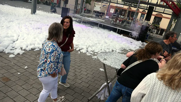 Zwei Frauen unterhalten sich auf einem gepflasterten Platz neben einer großen Menge weißen Schaums vor einer Bühne.