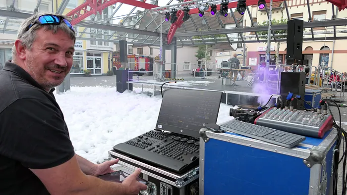 DJ Hansi lächelt an seinem DJ-Equipment mit Laptop und Mischpult vor einer Bühne, die mit weißem Schaum bedeckt ist.