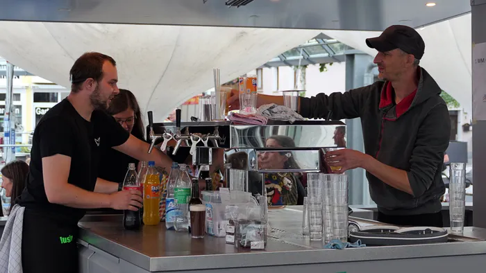 Zwei Männer arbeiten hinter einer Bar mit verschiedenen Flaschen, Gläsern und Zapfanlagen.