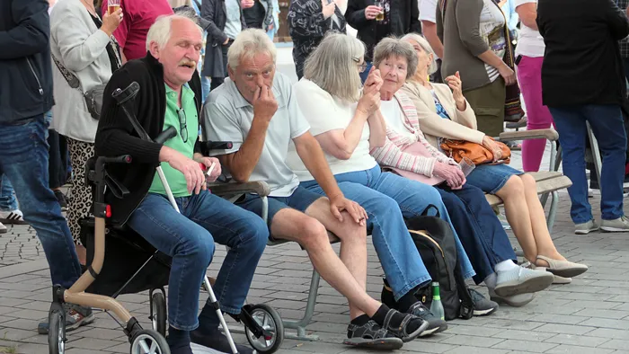 Eine Gruppe älterer Menschen sitzt auf Bänken im Freien, ein Mann nutzt einen Rollator.