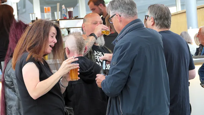 Eine Frau mit langen braunen Haaren hält ein Glas Bier und eine Zigarette, während im Hintergrund Gäste bei der Roxy Revival Party stehen.
