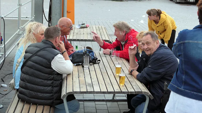 Eine Gruppe von Menschen sitzt an einem Holztisch im Freien und unterhält sich bei Getränken.