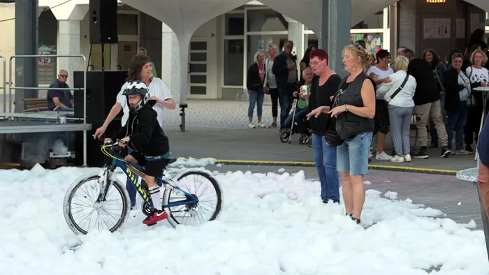 Ein Kind fährt mit dem Fahrrad durch eine dicke Schicht weißen Schaums auf einem Platz vor einer Gruppe von Menschen.