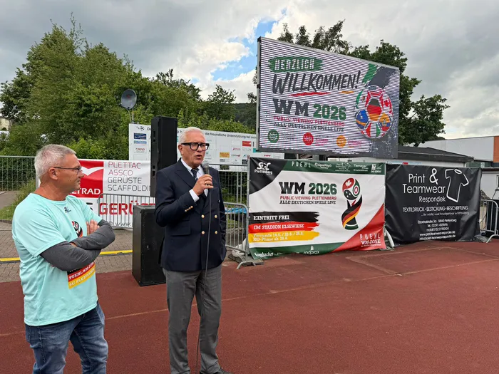 Ein Mann in einem orangefarbenen T-Shirt interviewt einen Mann in einem dunklen Anzug vor einer großen Leinwand, auf der „WM 2026“ steht. Im Hintergrund sind weitere Menschen und Werbebanner zu sehen.
