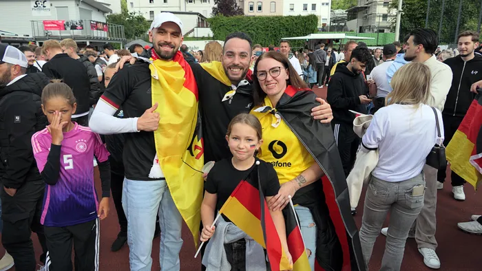 Eine Gruppe von Menschen, darunter Kinder und Erwachsene, posiert lächelnd mit Deutschlandflaggen und in Deutschlandtrikots. Im Hintergrund sind weitere Fans und Gebäude zu sehen.