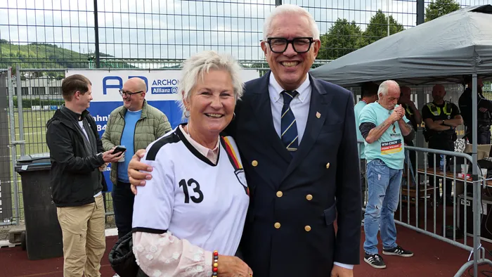 Zwei lächelnde ältere Menschen, eine Frau in einem Fußballtrikot mit der Nummer 13 und ein Mann im Anzug, stehen eng umschlungen vor einer Absperrung. Im Hintergrund sind weitere Zuschauer und ein Stadion zu sehen.