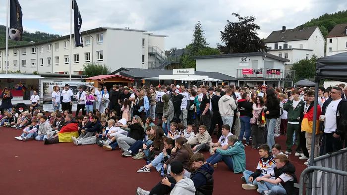 Eine große Menschenmenge sitzt und steht auf einem Platz vor einer Reihe von Gebäuden und verfolgt ein Fußballspiel. Viele Fans schwenken Deutschlandflaggen und jubeln über den Sieg der Nationalmannschaft.