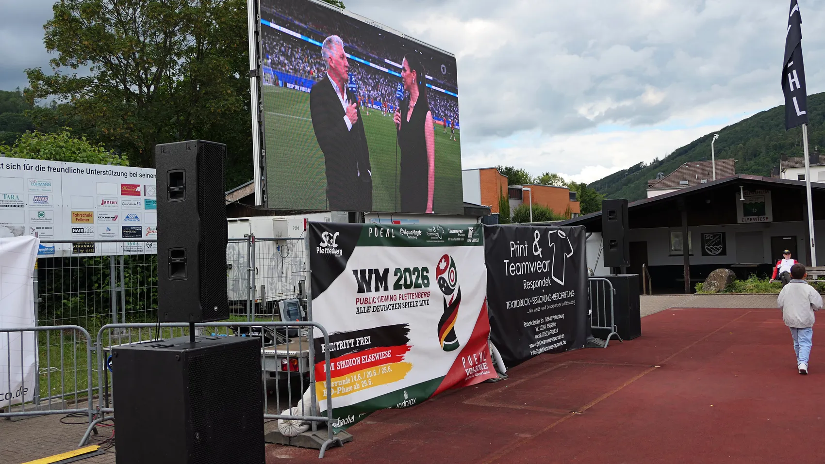 Große Videoleinwand zeigt Fußballspiel, davor eine Absperrung und Banner mit der Aufschrift