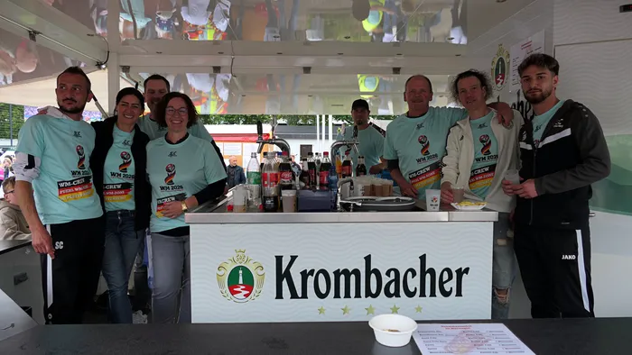 Sechs Personen stehen hinter einer Theke mit Getränken und Snacks. Sie tragen alle T-Shirts mit der Aufschrift 
