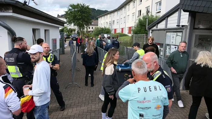 Polizeibeamte und Sicherheitskräfte kontrollieren den Zugang zu einer Menschenmenge vor einer Absperrung. Im Hintergrund sind Gebäude und Bäume zu sehen. Die Fans sind anlässlich des WM-Spiels Deutschland gegen Curacao zusammengekommen.