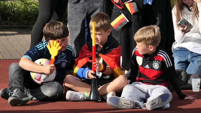 Drei Jungen sitzen auf dem Boden und beschäftigen sich mit Fußball-Utensilien. Links hält ein Junge einen Fußball und trägt Torwarthandschuhe, in der Mitte bläst ein anderer in eine Tröte, und rechts sitzt ein Junge mit einem Fußballtrikot. Im Hintergrund sind jubelnde Menschen und Deutschlandflaggen zu sehen.
