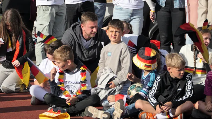 Eine Gruppe von Menschen, darunter viele Kinder, sitzt und schaut aufgeregt zu. Einige halten Deutschlandflaggen hoch und tragen Fanartikel. Im Hintergrund sind weitere Zuschauer zu sehen.