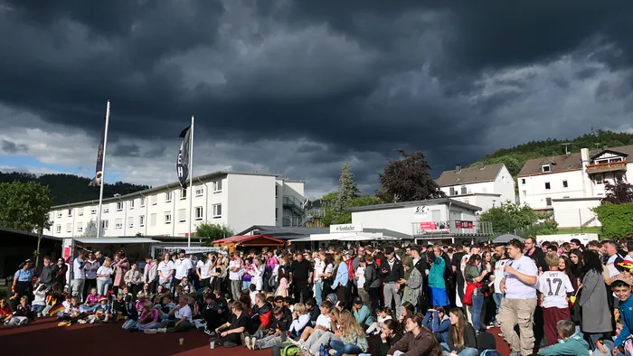 Eine große Menschenmenge jubelt vor einer modernen Gebäudeanlage, im Hintergrund eine bewaldete Hügellandschaft unter einem dramatischen Himmel. Viele Zuschauer halten Deutschlandfahnen hoch und feiern den Sieg der Nationalmannschaft.
