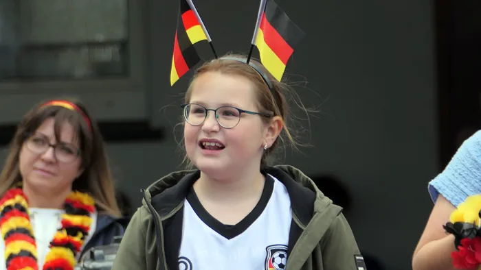 Junge Frau mit Brille und Deutschland-Trikot jubelt in einer Menschenmenge. Sie trägt zwei kleine Deutschlandflaggen auf einem Stirnband. Im Hintergrund sind weitere Fans mit Deutschlandfarben zu sehen.
