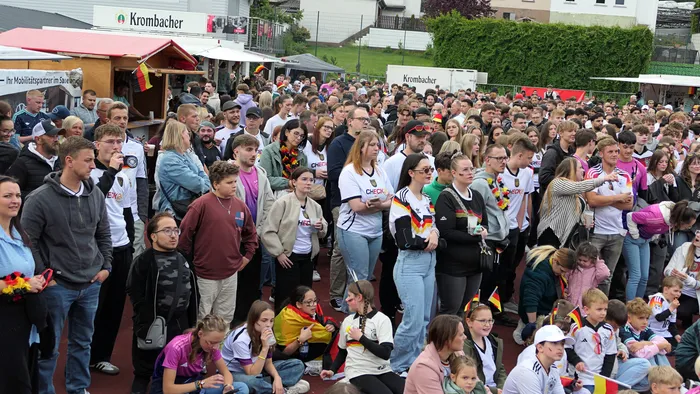 Eine große Menschenmenge jubelt und schaut auf eine Leinwand, viele tragen Deutschland-Trikots und schwenken Deutschland-Flaggen. Im Hintergrund sind Buden und weitere Fans zu sehen.