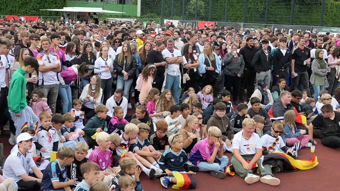 Eine große Menschenmenge jubelt und schaut auf eine nicht sichtbare Leinwand. Viele Fans tragen Trikots der deutschen Nationalmannschaft und schwenken Deutschlandflaggen.