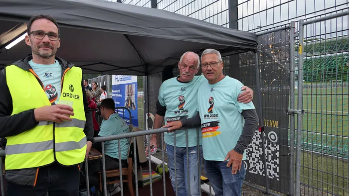 Drei Männer stehen vor einer Metallabsperrung, im Hintergrund eine Fan-Arena mit Zuschauern und einer großen Leinwand. Der Mann links trägt eine Warnweste mit der Aufschrift 