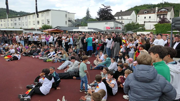 Eine große Menschenmenge sitzt und steht auf einem roten Sportplatz und schaut in eine Richtung. Viele tragen Trikots der deutschen Nationalmannschaft, im Hintergrund sind Gebäude und Bäume zu sehen.