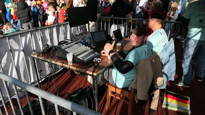 Ein Mann sitzt an einem Tisch mit Audio-Equipment und hält einen Pappbecher in der Hand. Im Hintergrund sind jubelnde Menschen und eine Deutschlandflagge zu sehen.