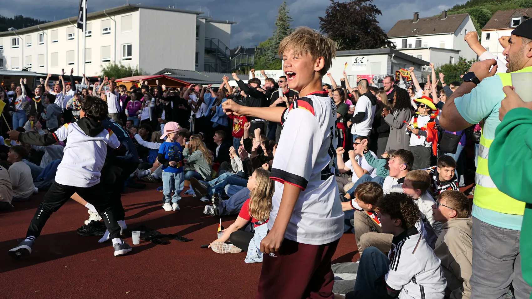 Jubelnde Menschenmenge vor Wohnhäusern, viele in Deutschland-Trikots, feiern den Sieg der Nationalmannschaft bei der Fußball-WM. Ein Junge im Vordergrund gestikuliert aufgeregt.