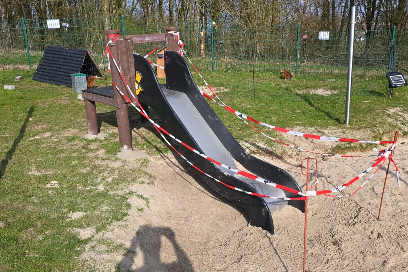 Abgesperrte Rutsche auf einem Spielplatz, umgeben von Sand und Gras. Im Hintergrund sind Hühner und ein Zaun zu sehen.