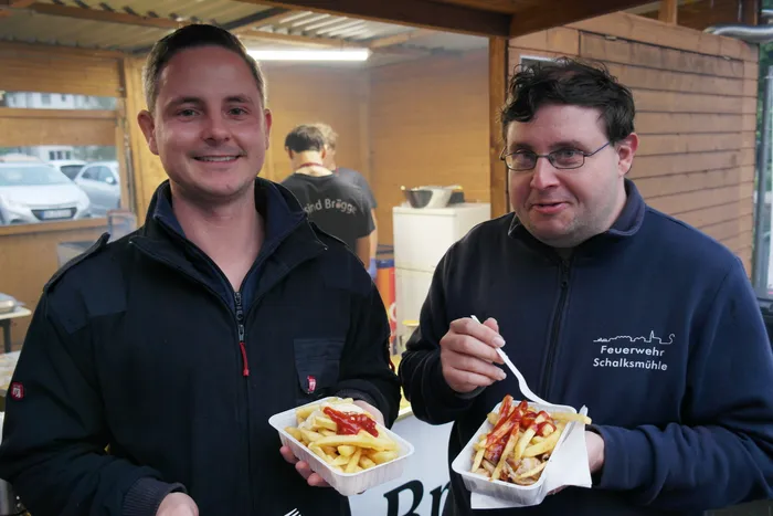 Zwei Männer lächeln in die Kamera und halten Schalen mit Pommes Frites und Soße. Im Hintergrund steht ein Imbisswagen und sind weitere Personen zu sehen.