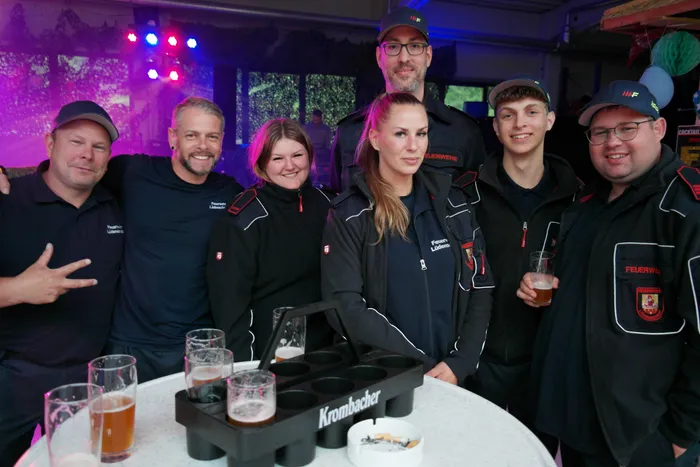 Eine Gruppe von Feuerwehrleuten in Uniformen steht lächelnd vor einer Bar und hält Biergläser. Auf dem Tisch stehen weitere Getränke und ein Aschenbecher mit Zigaretten.