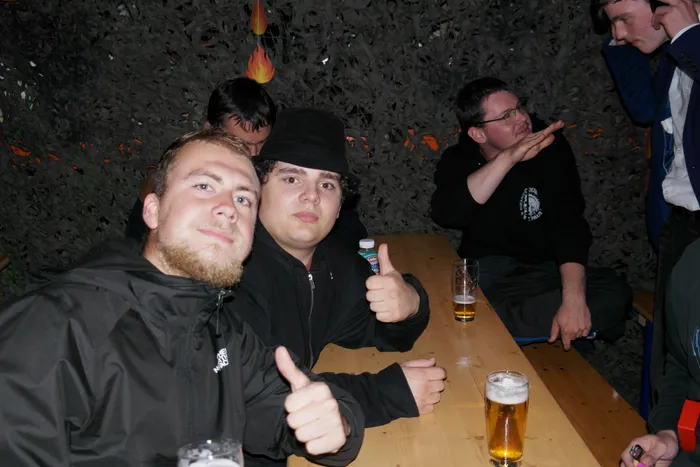 Zwei junge Männer lächeln in die Kamera und zeigen Daumen hoch, während sie an einem Tisch mit Biergläsern sitzen. Im Hintergrund sind weitere Personen und eine dekorative Wand zu sehen.