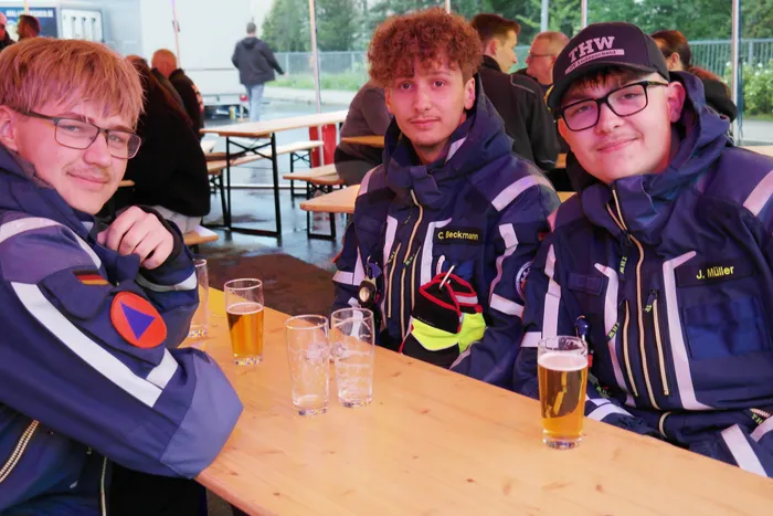 Drei THW-Helfer in blauen Einsatzjacken sitzen an einem Biertisch und halten Biergläser in der Hand. Im Hintergrund sind weitere Personen und Zelte auf einem Feuerwehrfest zu sehen.