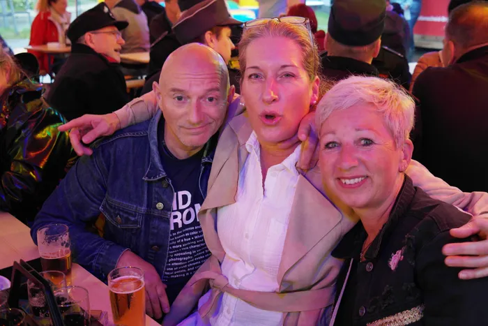 Drei lächelnde Frauen und ein Mann umarmen sich vor einer Menschenmenge auf einem Feuerwehrfest. Der Mann hält ein Bierglas in der Hand.
