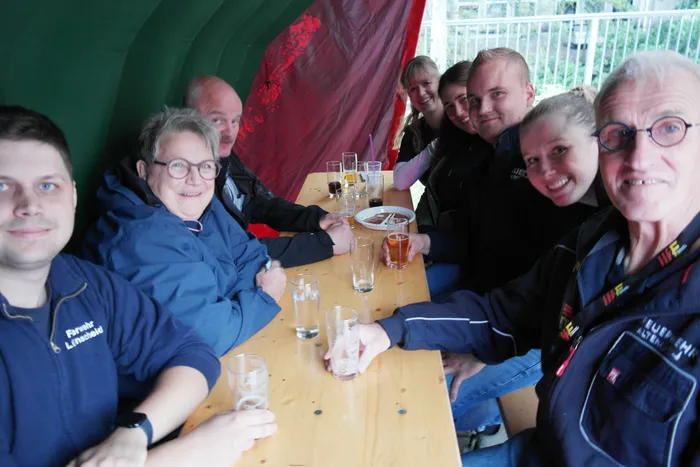 Eine Gruppe von Feuerwehrleuten und Gästen sitzt an einem Holztisch in einem Zelt und genießt Getränke. Einige lächeln in die Kamera.