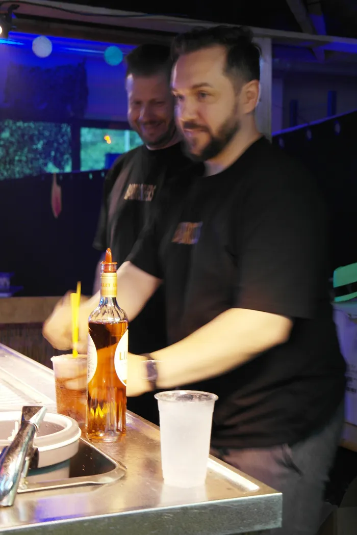 Zwei Männer hinter einer Bar schenken Getränke ein, im Hintergrund blaue Beleuchtung und weitere Personen.