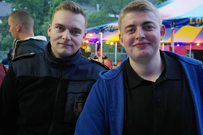 Zwei junge Feuerwehrleute in Uniformen lächeln in die Kamera, im Hintergrund ein Fest mit Lichtern und Zelten.