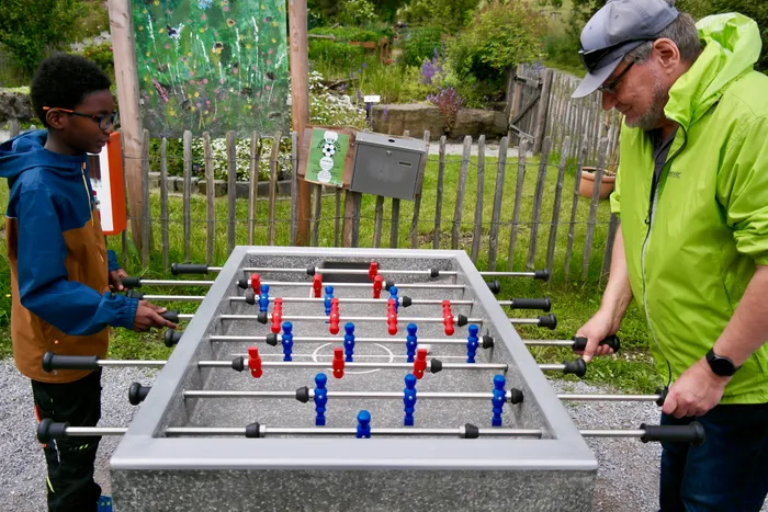 Ein Junge und ein Mann spielen Tischfußball im Freien. Im Hintergrund ist ein Garten mit einer bunten Wandgestaltung zu sehen.