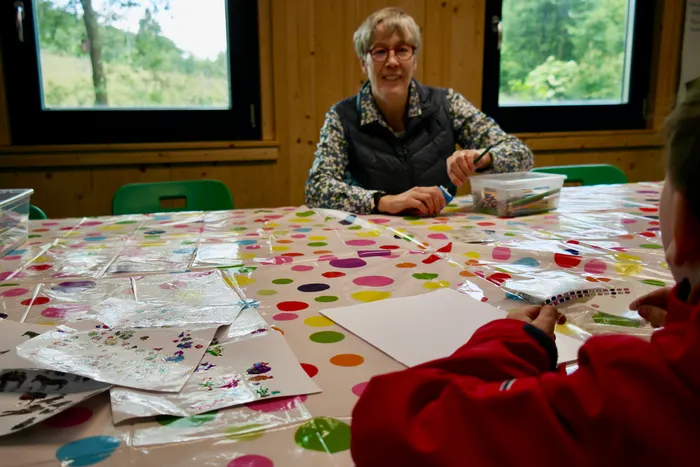 Eine Frau sitzt an einem mit farbigen Punkten versehenen Tisch und hält einen Stift. Vor ihr liegen Bastelmaterialien und Papier, während im Hintergrund eine Person in roter Jacke zu sehen ist.