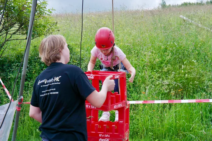 Ein Junge hilft einem Mädchen mit einem Helm, auf eine aus Coca-Cola-Kisten gebaute Plattform zu klettern. Im Hintergrund eine Wiese und Büsche.