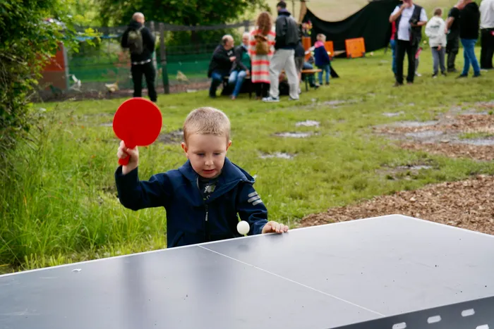 Ein Junge spielt Tischtennis im Freien. Er hält einen Schläger hoch und bereitet sich auf einen Schlag vor, während der Ball auf der Tischtennisplatte liegt. Im Hintergrund sind weitere Menschen und Zelte zu sehen.