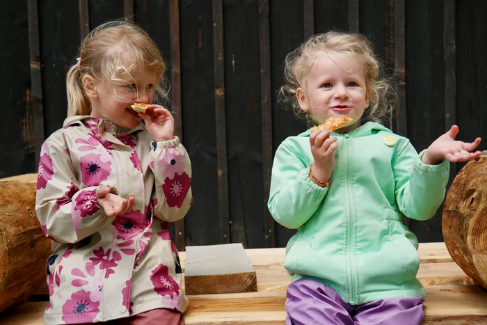 Zwei kleine Mädchen essen Pizza. Beide tragen bunte Jacken und schauen in die Kamera.