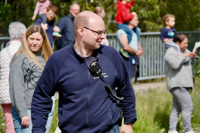 Ein Mann mit Glatze und Brille trägt eine blaue Jacke mit dem Aufdruck 