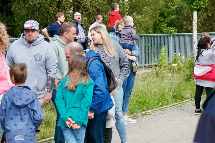 Menschengruppen stehen und gehen auf einem Weg, einige Kinder halten Hände. Im Hintergrund ist ein grauer Zaun und Grün zu sehen.