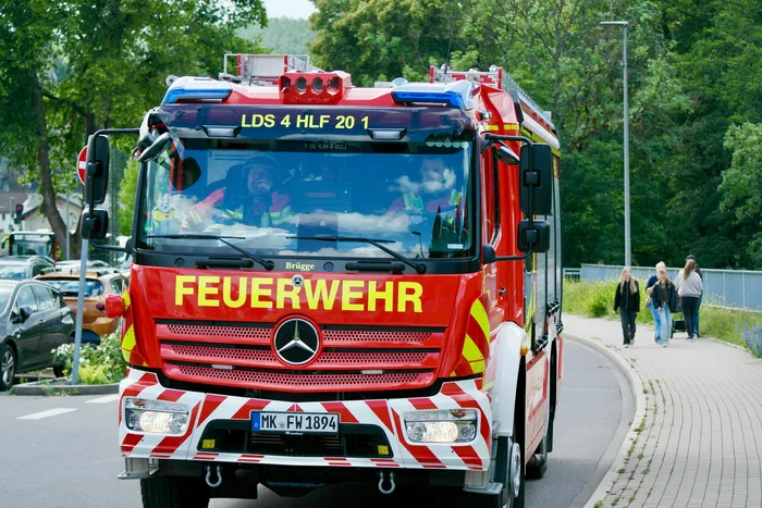 Roter Feuerwehrauto mit der Aufschrift „FEUERWEHR“ steht auf einer Straße, während im Hintergrund Menschen vorbeigehen. Zwei Feuerwehrleute sind im Fahrhaus zu sehen.