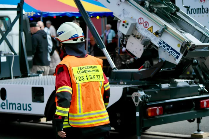 Ein Feuerwehrmann mit Helm und Warnweste steht vor einem großen, weißen Kranfahrzeug mit der Aufschrift „Böcker“. Auf der Weste ist „Feuerwehr Lüdenscheid“ und eine Kennnummer zu lesen. Im Hintergrund sind Menschen und ein buntes Zelt zu erkennen.