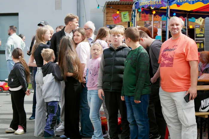 Menschen stehen vor bunten Buden auf einem Volksfest. Kinder und Erwachsene warten in einer Schlange, einige schauen in Richtung der Buden.